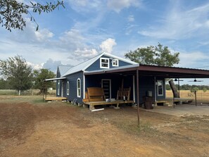 Ein einstöckiges Haus mit einer Veranda und einem überdachten Bereich, umgeben von trockenem Gras und Bäumen.