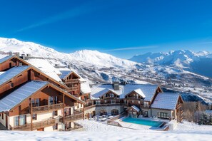 Ein Chalet-Hotel mit Schwimmbad, Holzbalkonen und Blick auf schneebedeckte Berge.