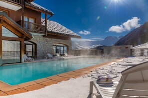 Ein Poolbereich mit Liegestühlen, ein Steinhaus und Blick auf schneebedeckte Berge.