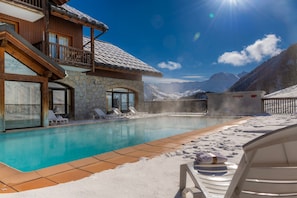A pool area with lounge chairs, a stone building, and a view of snow-capped mountains.