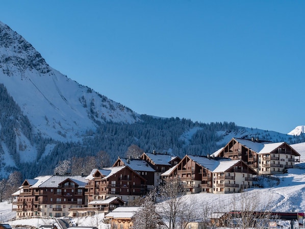 A cluster of chalet-style buildings with multiple balconies, set against a backdrop of snow-covered mountains and clear blue sky.