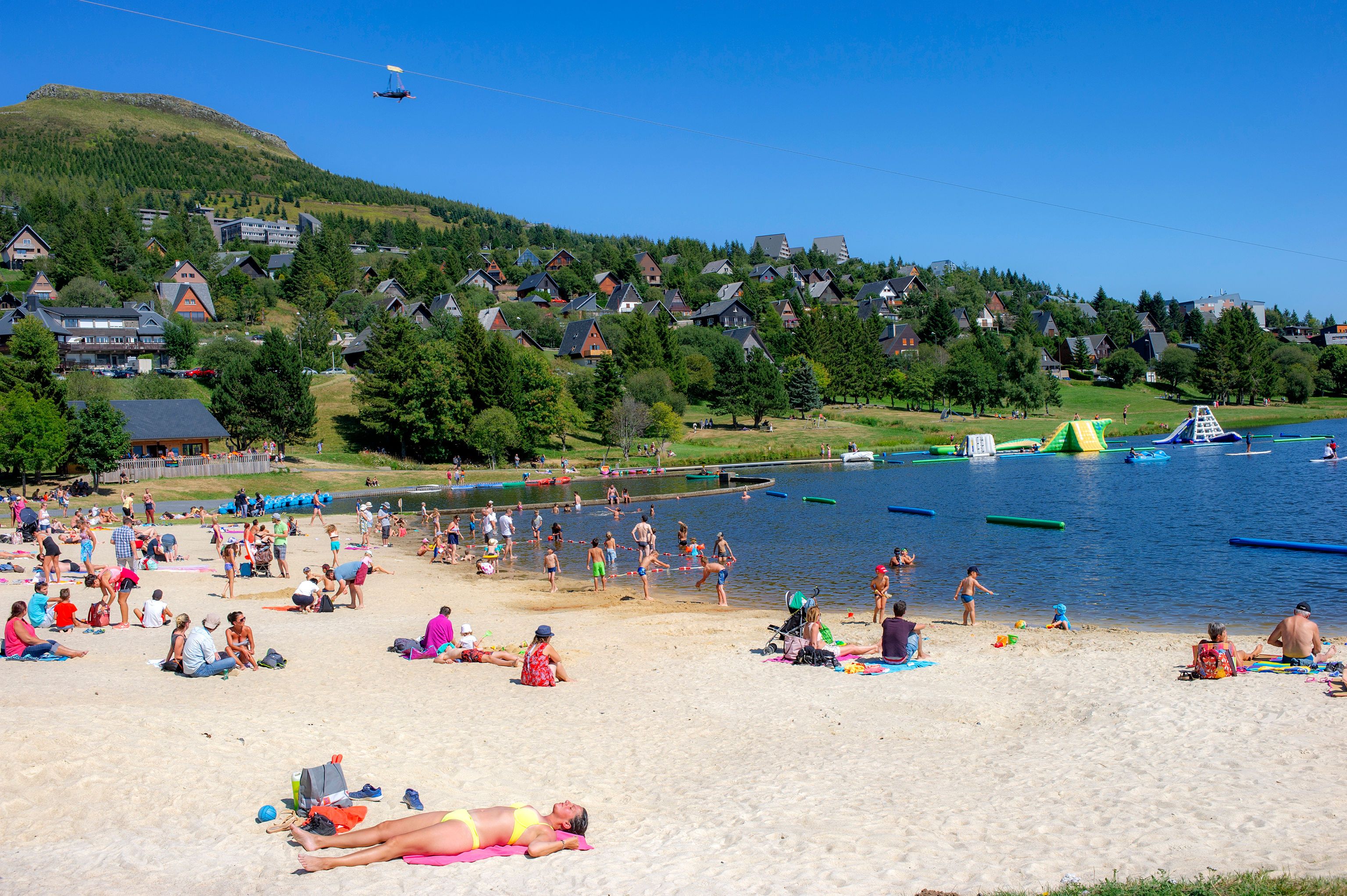 Een strand met mensen die zonnen en zwemmen, omgeven door huizen en een heuvel op de achtergrond.