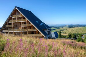 A large, multi-story wooden building with a sloped roof, surrounded by a field of purple flowers and a view of distant hills.
