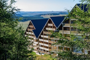 A multi-story wooden chalet with balconies, set against a mountainous backdrop.