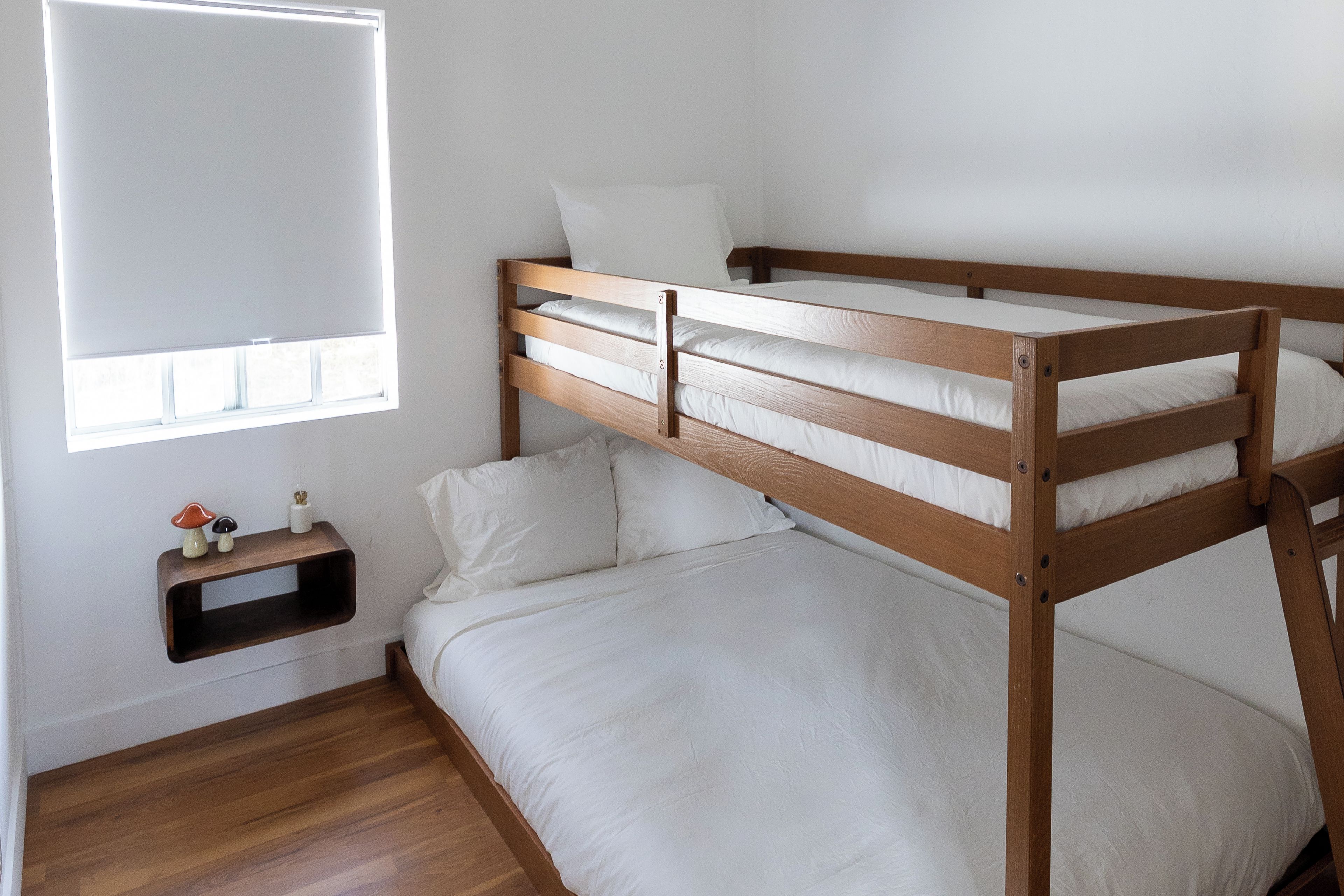 A bunk bed with a wooden frame and white bedding, a small shelf with decorative items, and a window with blinds.