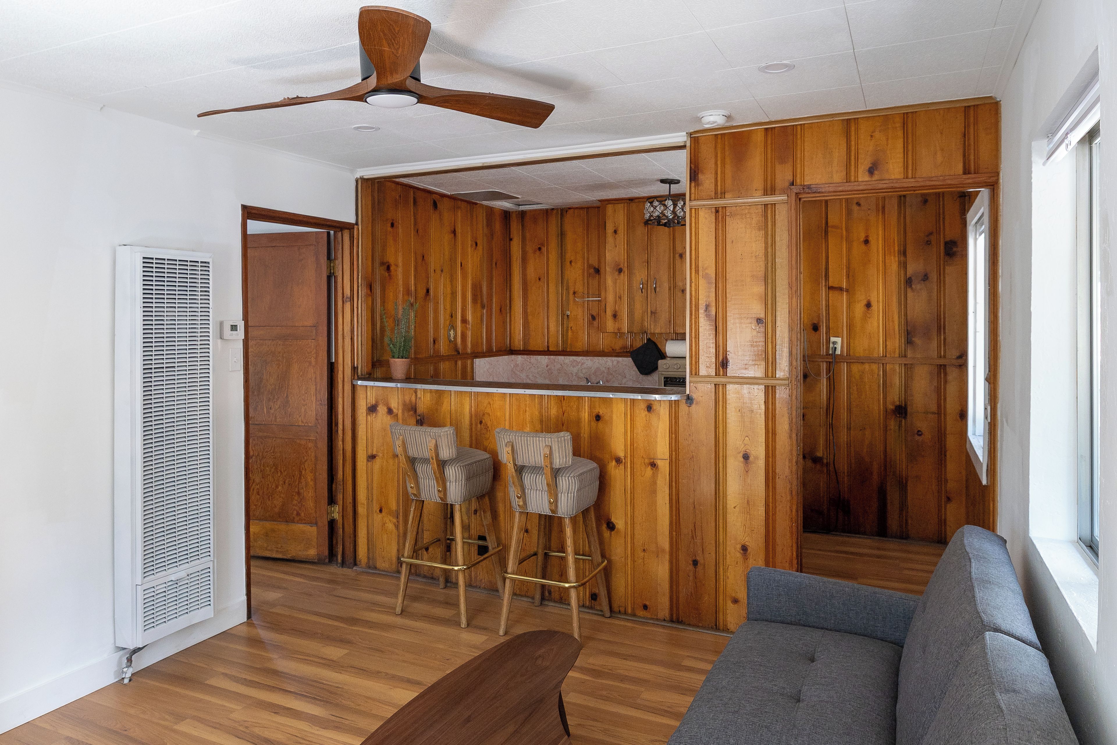 Ein Zimmer mit Holzwandverkleidung, einem Deckenventilator, einer Bar mit Hockern, einer Couch und einem Heizkörper.