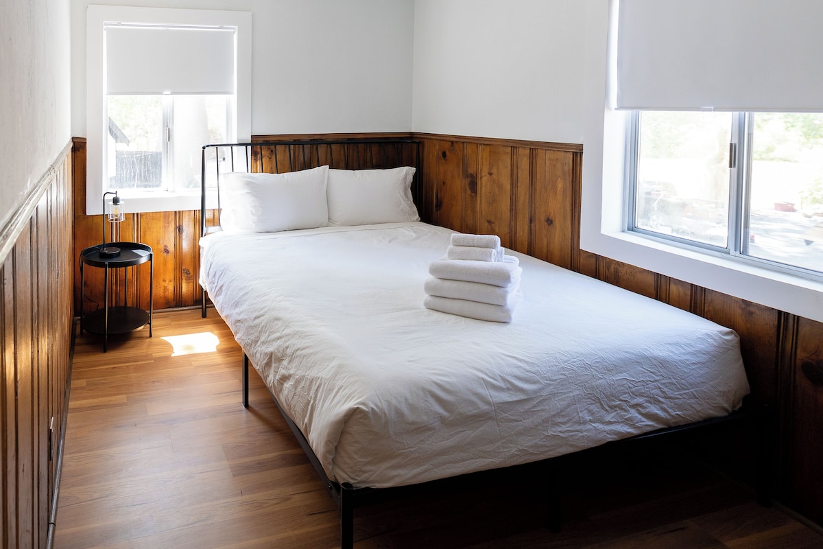 A single-bed room with a wooden headboard, a bedside table with a lamp, and a window with a view of the outside.