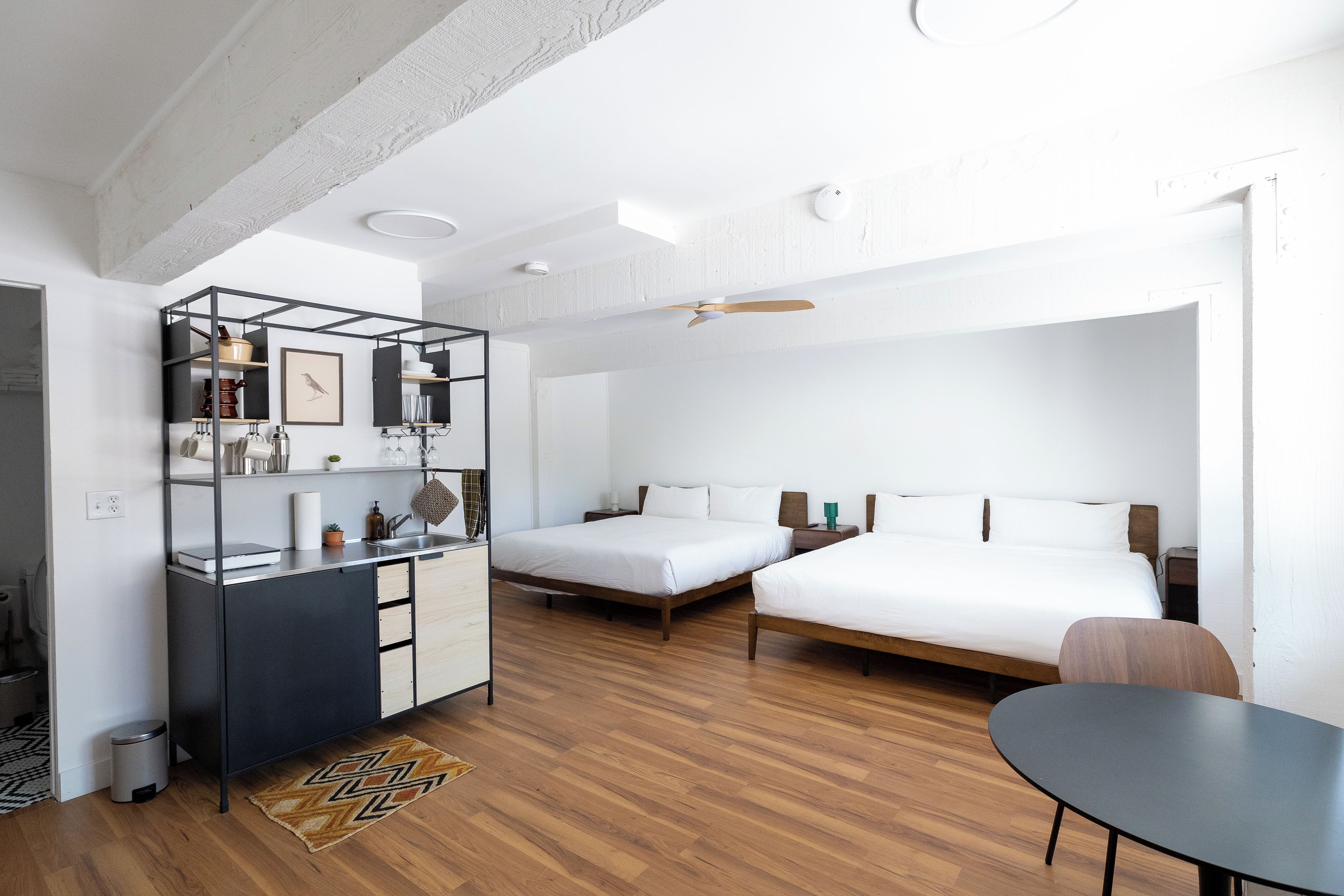 A modern hotel room with a kitchenette, two beds, a dining table, and a ceiling fan.