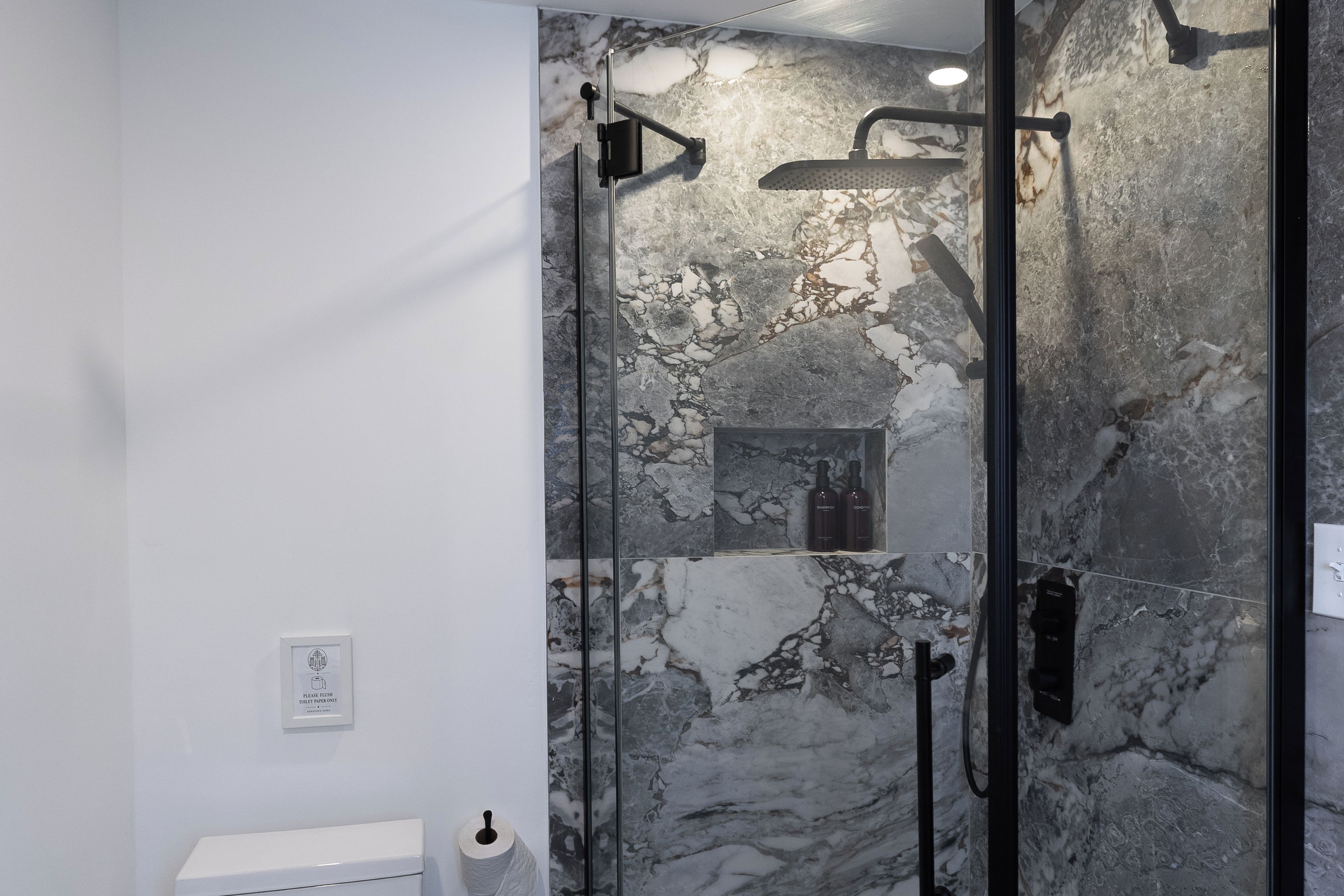 A modern bathroom with a glass-enclosed shower, marble walls, and a toilet paper dispenser.