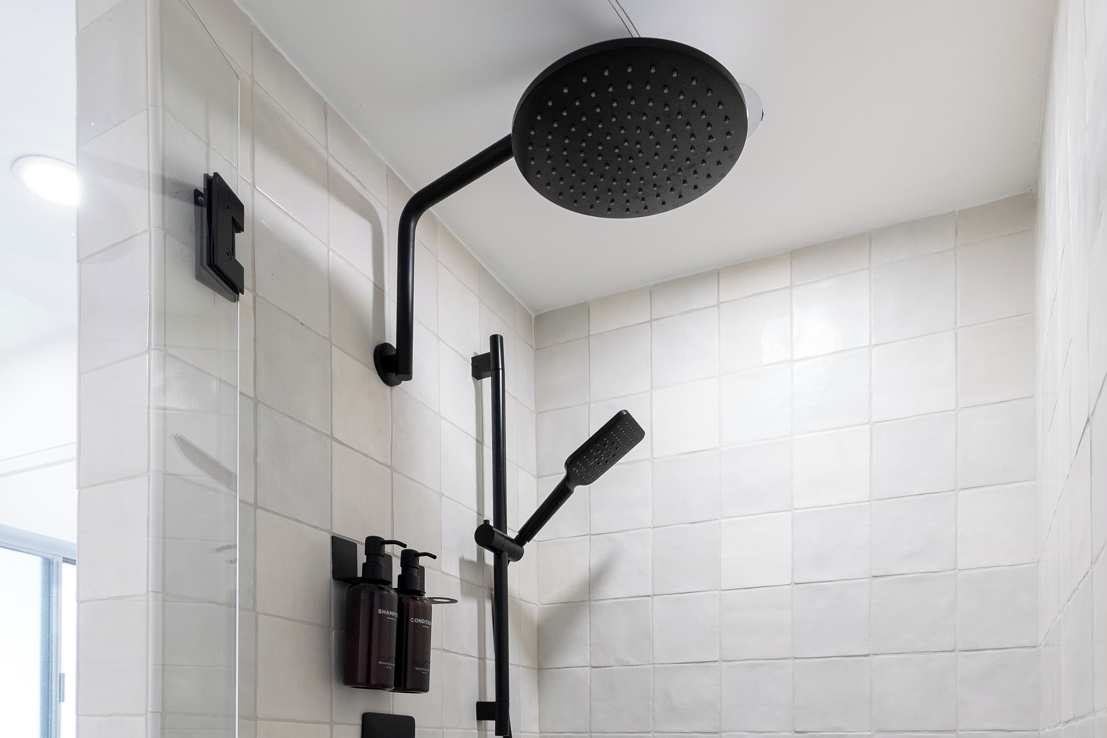Un baño moderno con paredes de azulejos blancos, un cabezal de ducha negro y un rociador de mano.