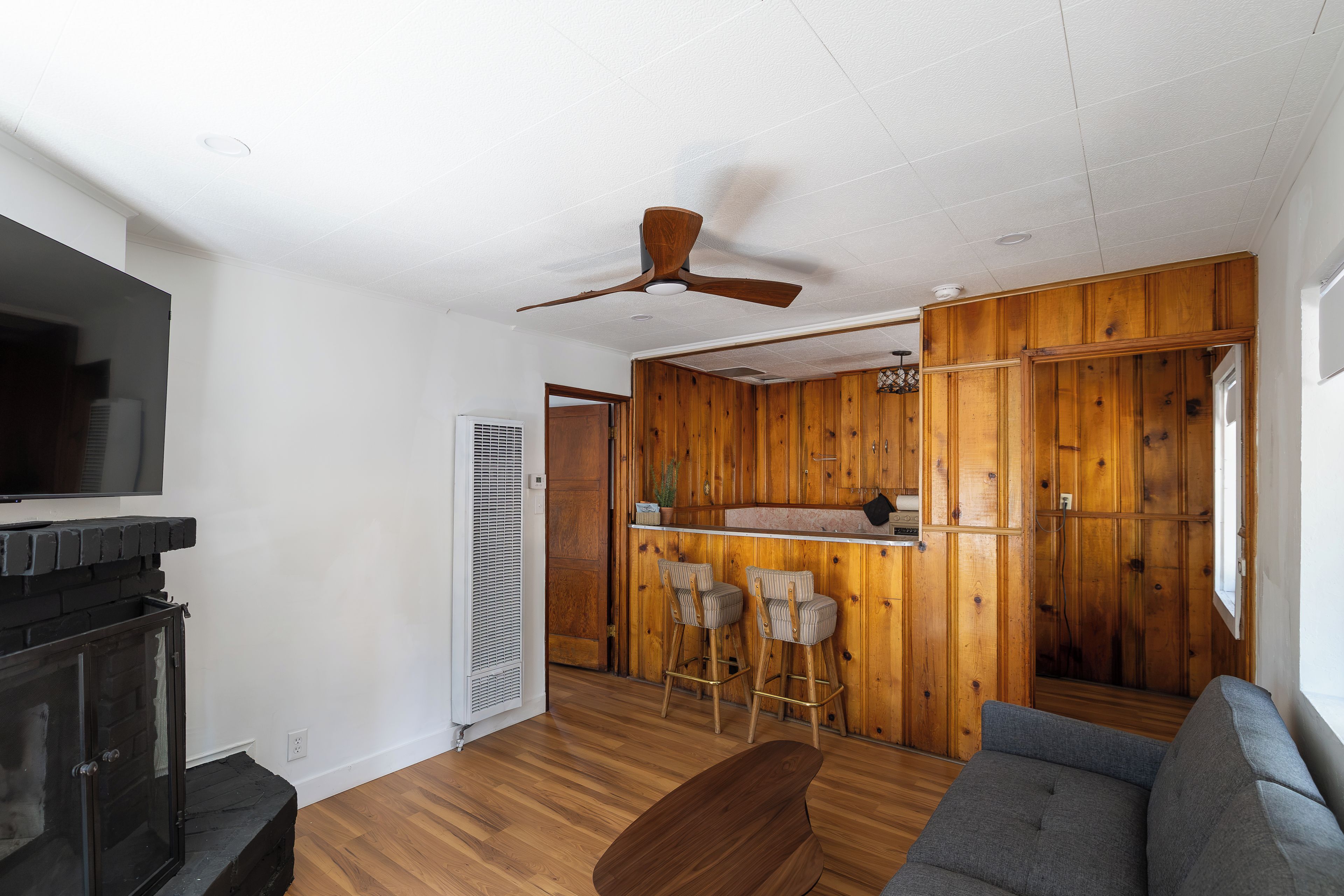 A living room with a fireplace, a ceiling fan, a sofa, and a kitchen area with wooden paneling.