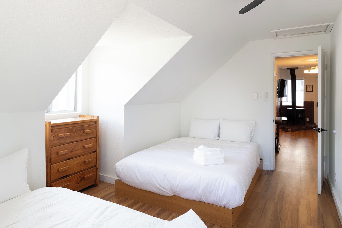 A bedroom with a sloped ceiling, a wooden dresser, a bed with white linens, and a view of a hallway.