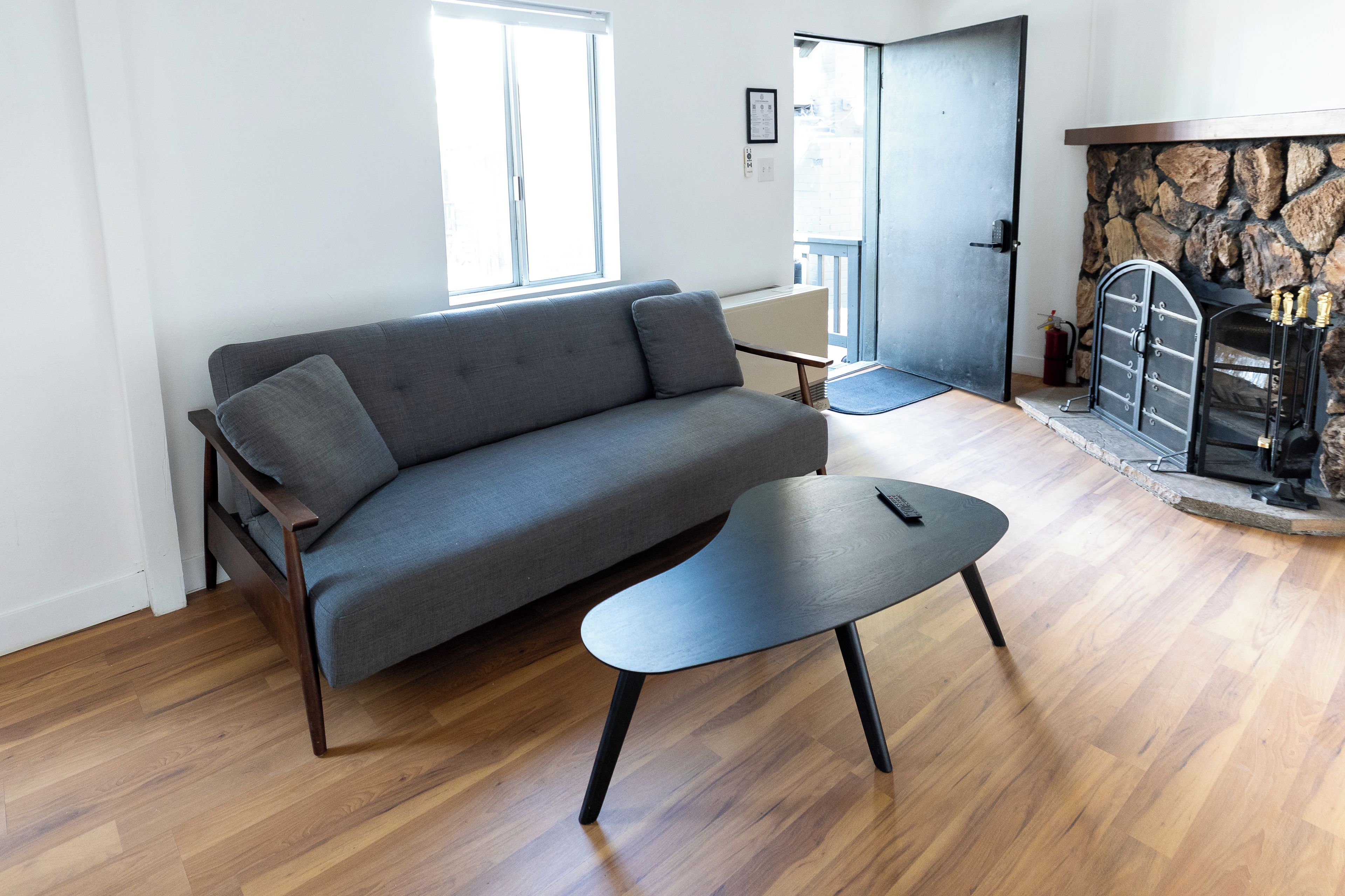 Ein modernes Wohnzimmer mit einem grauen Sofa, einem kleinen schwarzen Couchtisch, Holzböden, einem Kamin aus Stein und einer Tür zu einem anderen Raum.