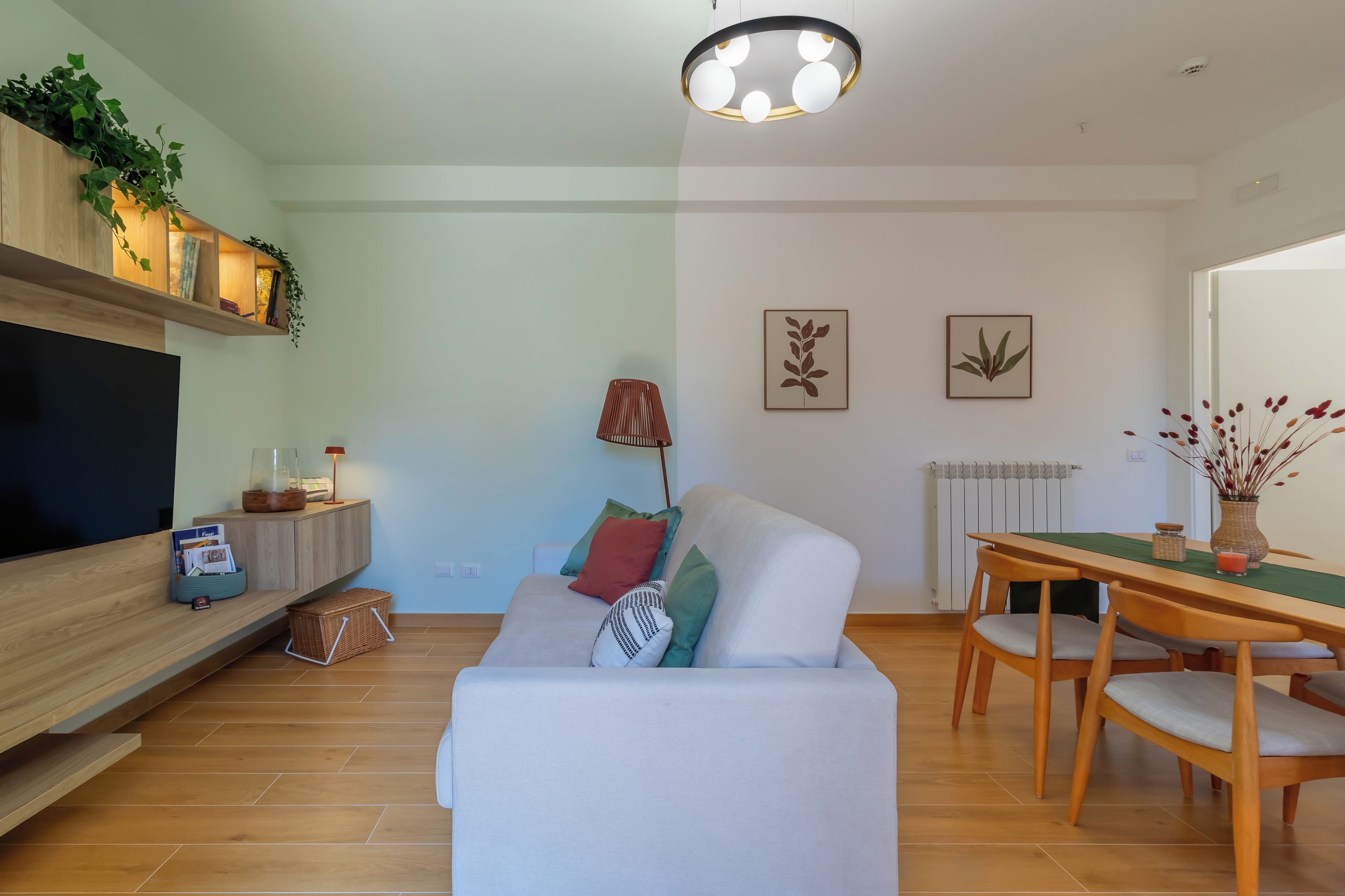 A modern living room with a flat-screen TV, a sofa, a dining table with chairs, and a ceiling light fixture.
