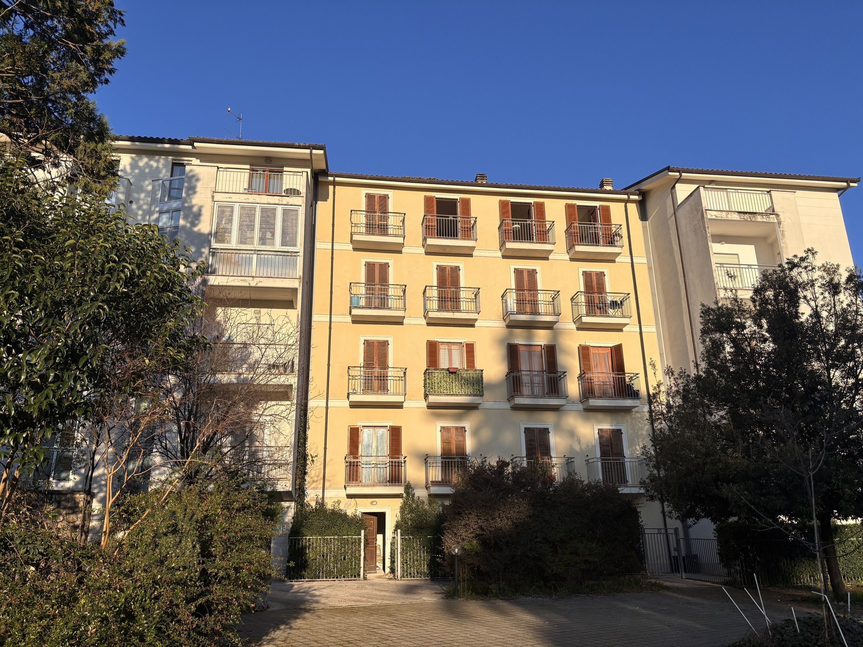 A multi-story residential building with balconies, a gated entrance, and surrounding greenery.