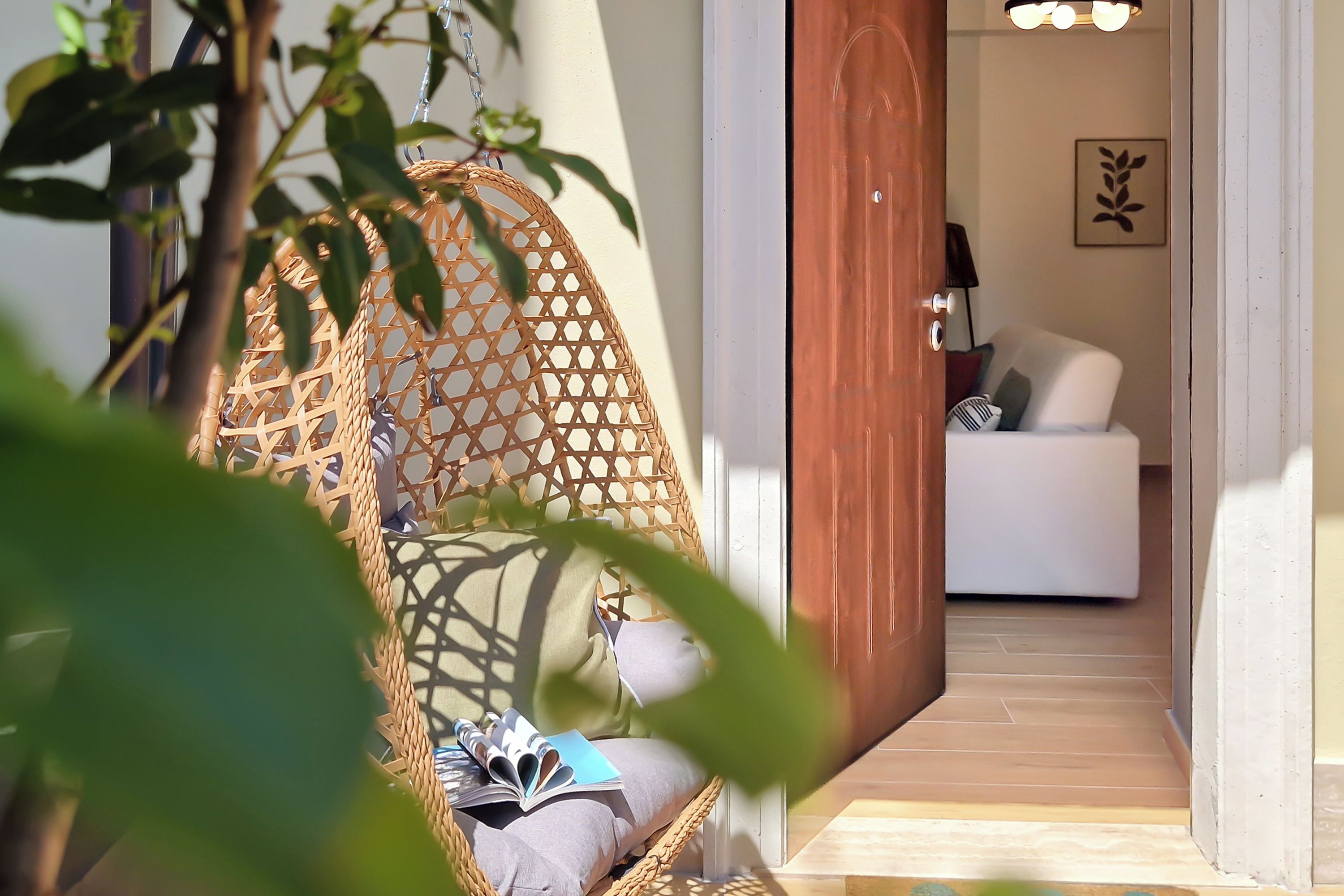 Una silla de mimbre con un cojín y un libro, un sofá blanco con almohadones, y una puerta que da a una habitación con una lámpara y un cuadro en la pared.