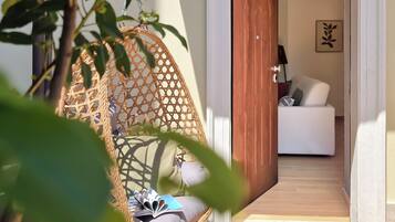 Una silla de mimbre con un cojín y un libro, un sofá blanco con almohadones, y una puerta que da a una habitación con una lámpara y un cuadro en la pared.