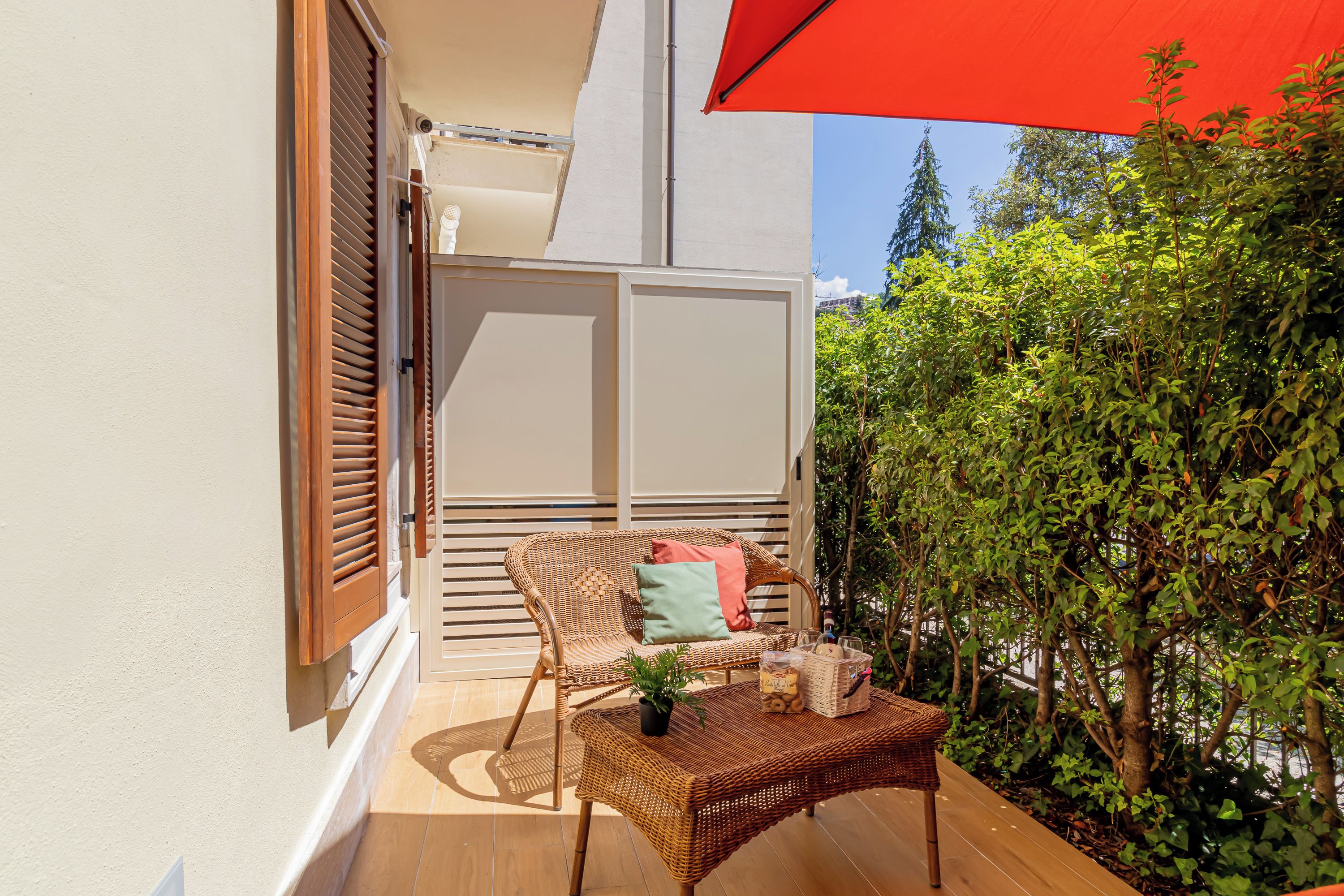 Un balcon avec une table et des chaises en osier, ainsi qu’un parasol rouge.