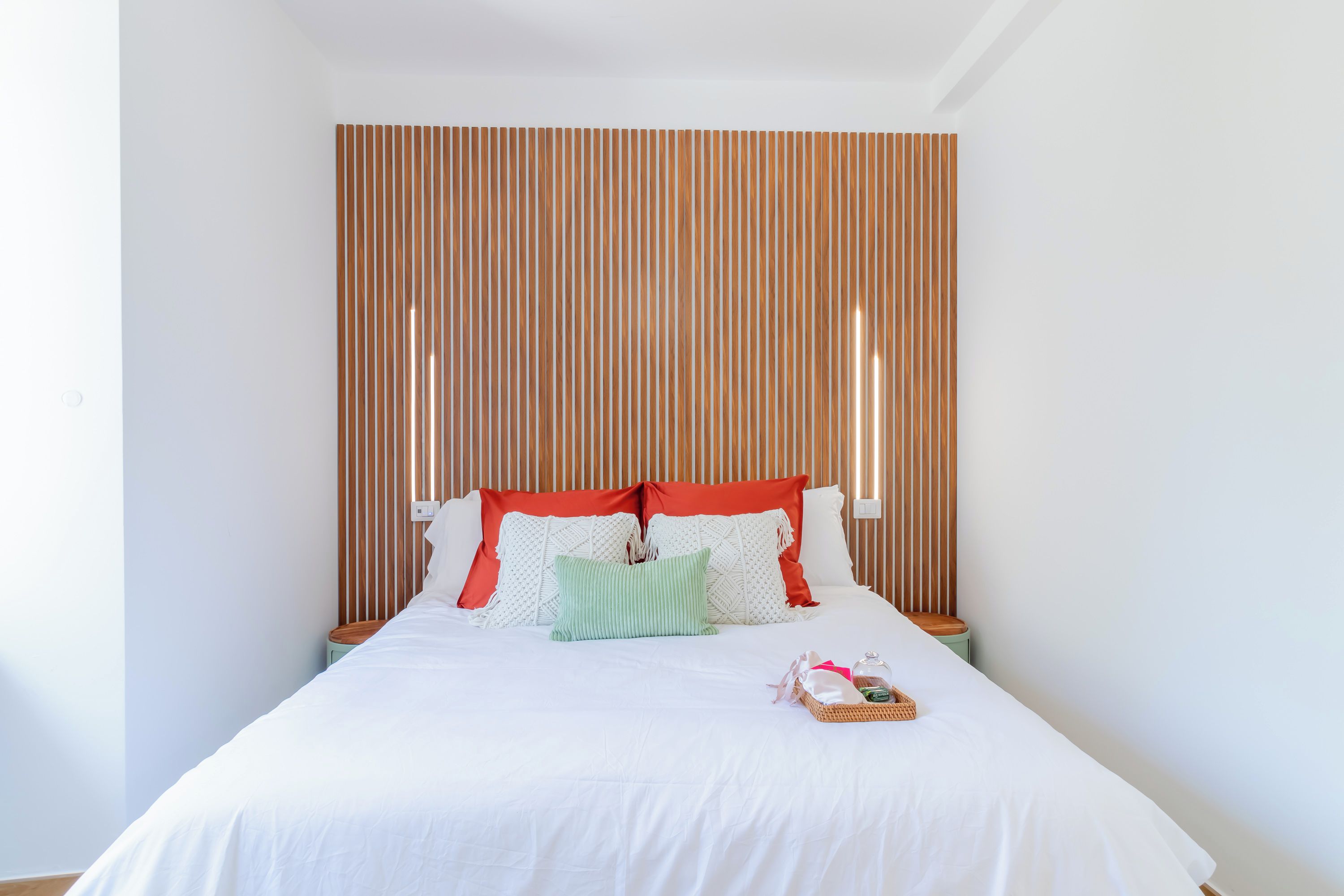 Una cama bien tendida con ropa de cama blanca y varias almohadas, una mesita de noche con una lámpara y un cabecero de madera con listones.