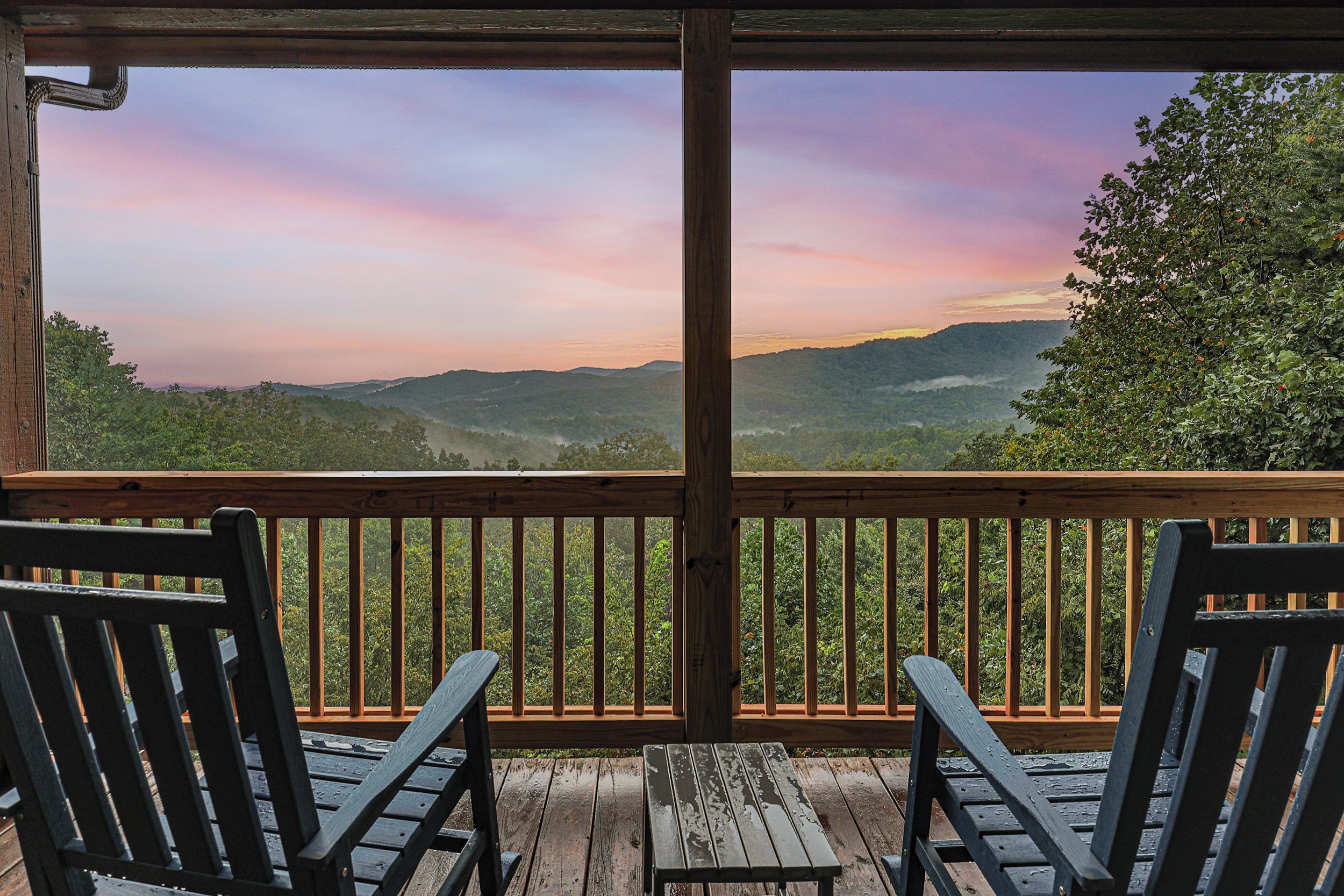 A wooden deck with two chairs and a table, overlooking a mountainous landscape with a sunset.