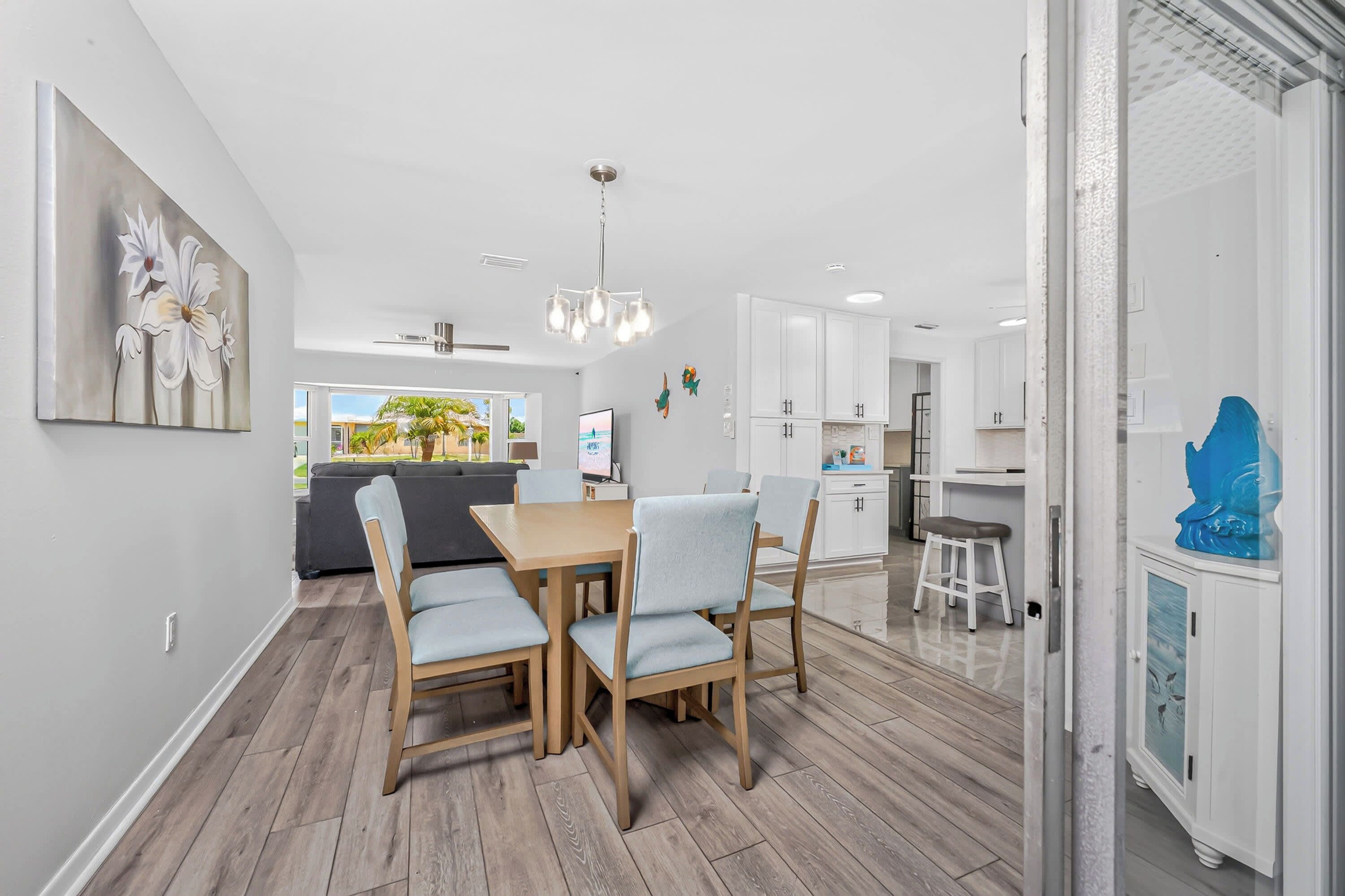 A modern kitchen with white cabinets, a dining area with a wooden table and chairs, and a view of the outdoors through a large window.