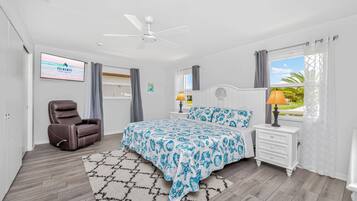 Ein Schlafzimmer mit einem Bett, einem Liegestuhl, einem Deckenventilator, einem Nachttisch mit Lampe und einem Fenster mit Vorhängen.