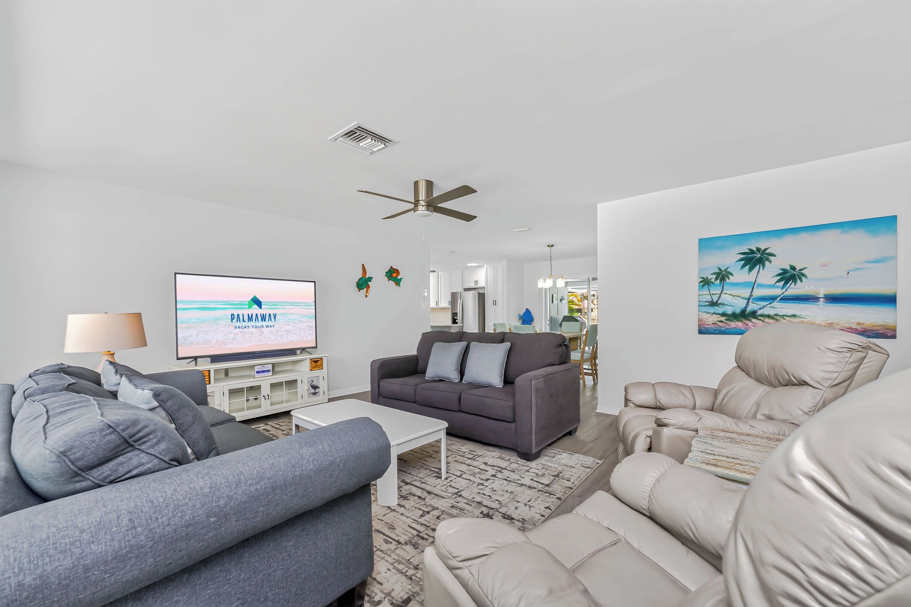 A modern living room with a flat-screen TV, grey sofas, a white coffee table, a ceiling fan, and a beach-themed painting on the wall.