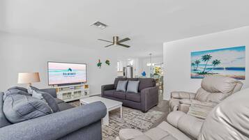 A modern living room with a flat-screen TV, grey sofas, a white coffee table, a ceiling fan, and a beach-themed painting on the wall.