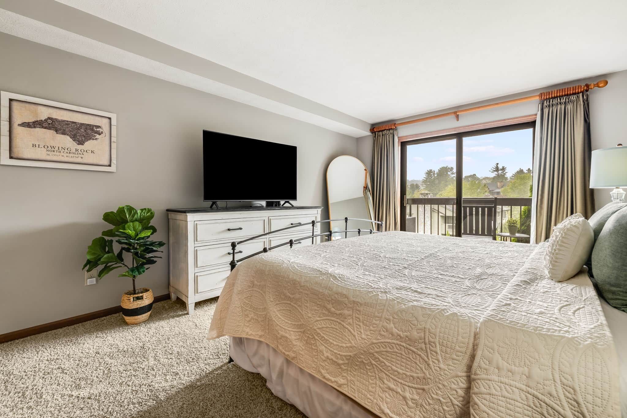 Un dormitorio con una cama grande, un televisor en una cómoda, una planta en maceta y un balcón con cortinas.
