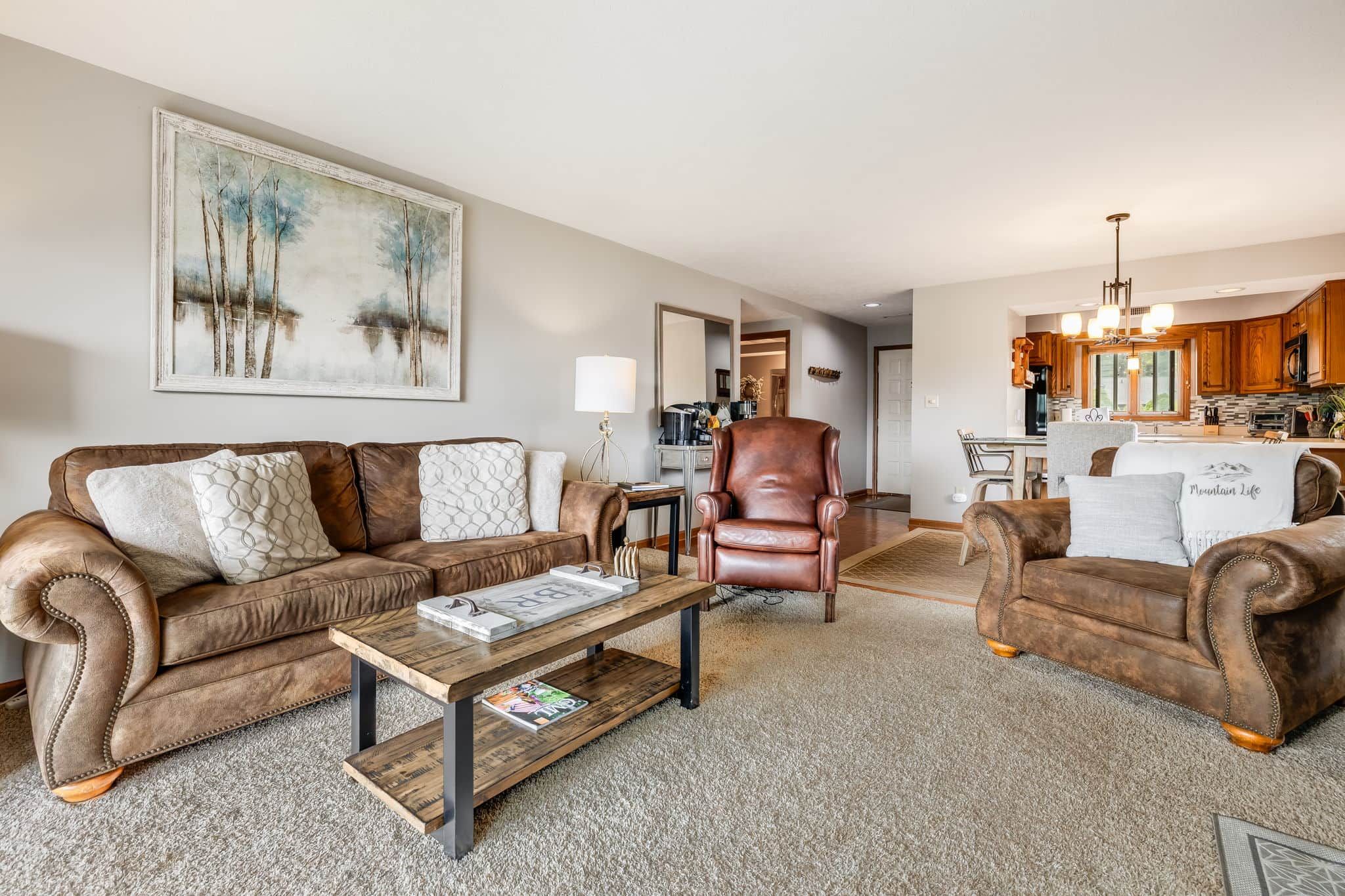 A living room with a brown leather sofa, a wooden coffee table, and a brown leather armchair. There is a painting on the wall and a kitchen in the background.