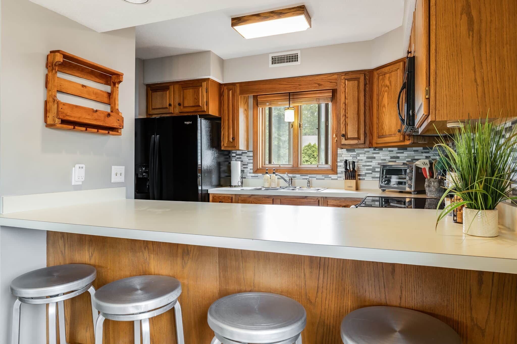 A kitchen with wooden cabinets, a black refrigerator, a large window, and a bar area with stools.