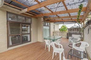 A covered patio area with a wooden deck, a glass table, white plastic chairs, and a hanging plant.