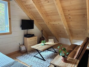 A cozy attic room with a wooden ceiling, a television, a guitar, a wooden desk, a couch, and a potted plant.