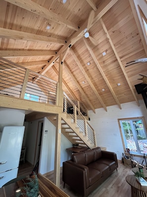 A spacious living area with a high ceiling, wooden beams, a staircase, a brown leather sofa, and a window providing a view of the outdoors.