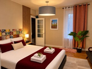 A hotel room with a bed, a bedside table with a lamp, a large window with curtains, a potted plant, and a wall with a colorful artwork. - ESCALE DE LA BREGERE (Limoges)