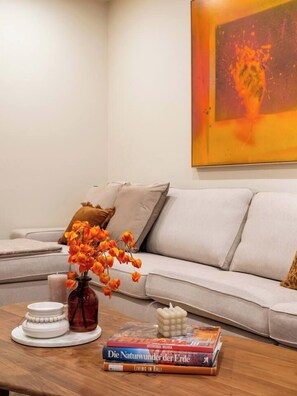 Uma sala de estar com um sofá, um vaso com flores laranja, uma vela e uma pilha de livros.