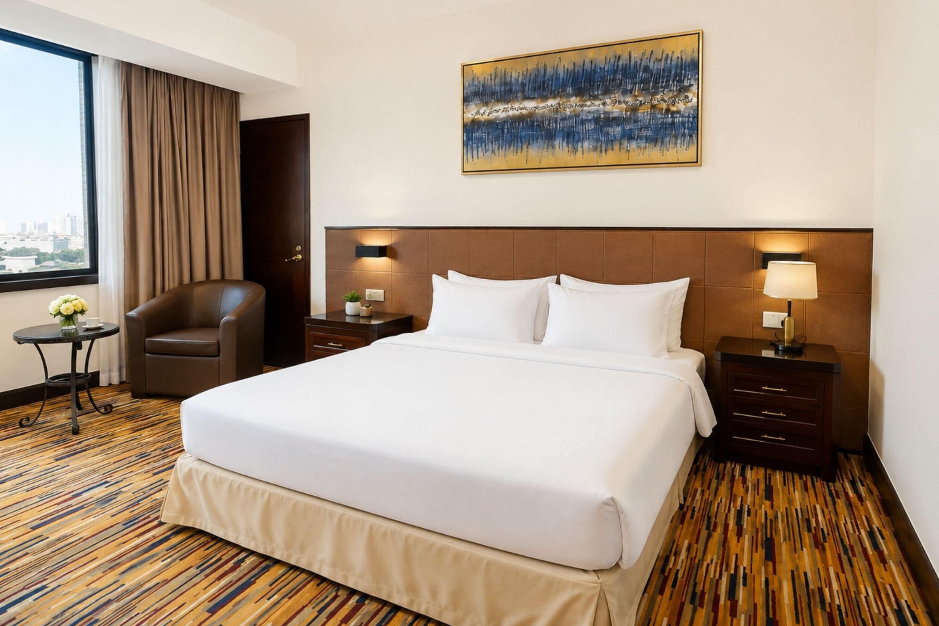 A hotel room with a large bed, a chair, a small table, and a painting on the wall.