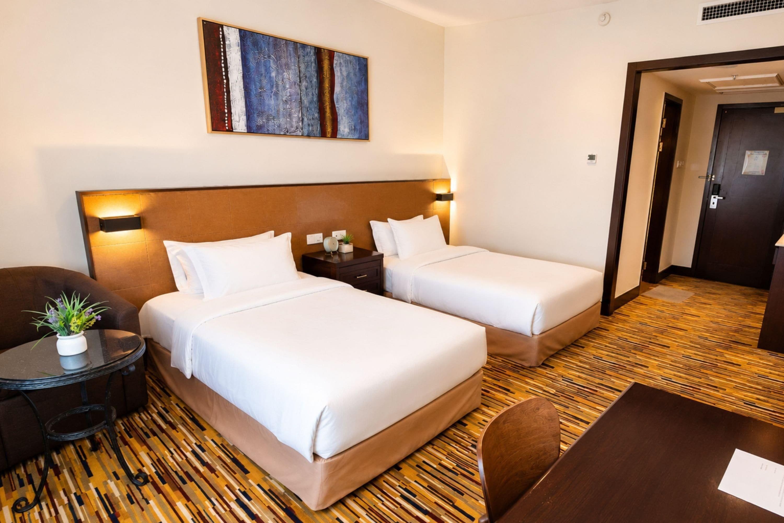 A hotel room with two beds, a small table, a chair, a nightstand, and a painting on the wall.