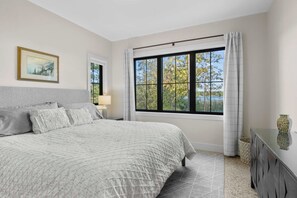 A bedroom with a large bed, a nightstand, a lamp, a window with curtains, and a painting on the wall. - Firefly Landing | Pet Friendly | Golf (Elk Rapids)