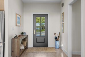 A hallway with a dark door, a wooden console table with decorative items, a blue vase with an umbrella, and framed pictures on the walls. - Firefly Landing | Pet Friendly | Golf (Elk Rapids)