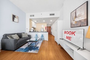 A modern living room with a grey sofa, a coffee table, and a rug. There is a kitchen area with a microwave and a sink. A sign on the wall reads 'BOOK ME'. - StayShort - Studio with best skyline view in JVT for 2! (JVT)