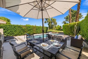 An outdoor patio with a pool, dining table, chairs, and a large umbrella. - Private Pool & Firepit Escape in Mission Hills Country Club | Mountain Views, Golf & Pickleball (Rancho Mirage)