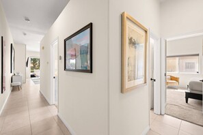 A hallway with framed artwork on the walls, a door leading to a living area, and a white chair in the distance. - Private Pool & Firepit Escape in Mission Hills Country Club | Mountain Views, Golf & Pickleball (Rancho Mirage)