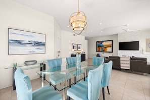 A modern dining area with a glass table and blue upholstered chairs, a chandelier, and artwork on the walls. - Private Pool & Firepit Escape in Mission Hills Country Club | Mountain Views, Golf & Pickleball (Rancho Mirage)