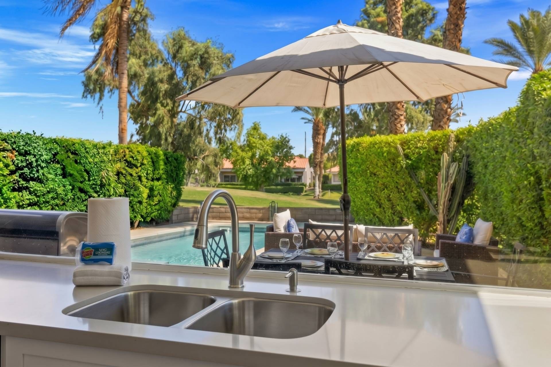 Une cuisine équipée d’un évier, un coin repas avec une table et des chaises, et une piscine en arrière-plan.