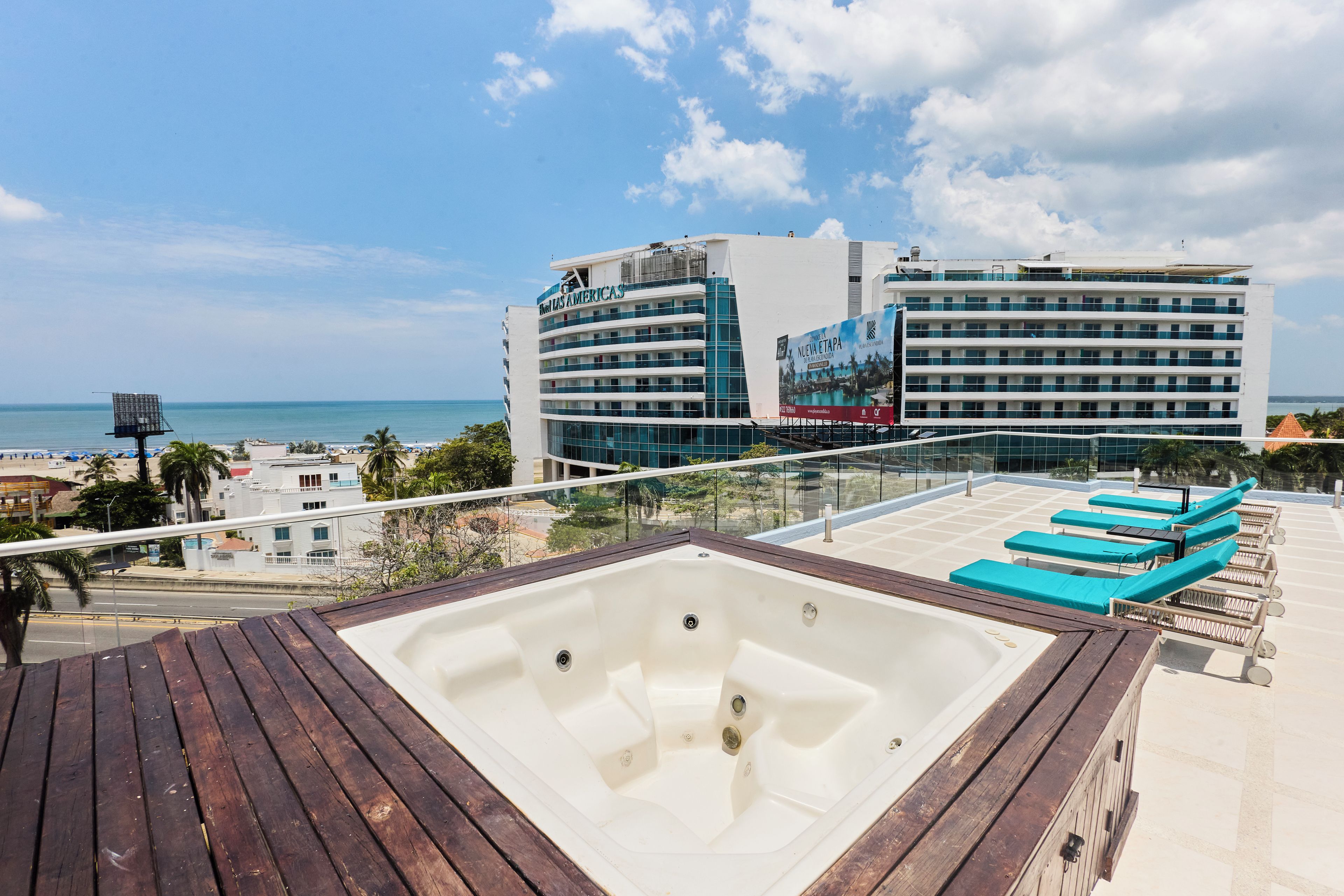 A rooftop pool area with a hot tub, lounge chairs, and a view of the ocean and hotel buildings.