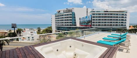 A rooftop pool area with a hot tub, lounge chairs, and a view of the ocean and hotel buildings.