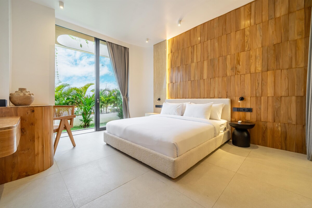 Quarto moderno com uma cama grande, um painel de parede de madeira, uma escrivaninha com cadeira e vista para o verde pela janela.