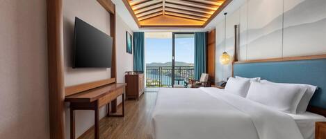 A hotel room with a large bed, a flat-screen TV, a wooden bench, a desk, and a view of the outdoors.