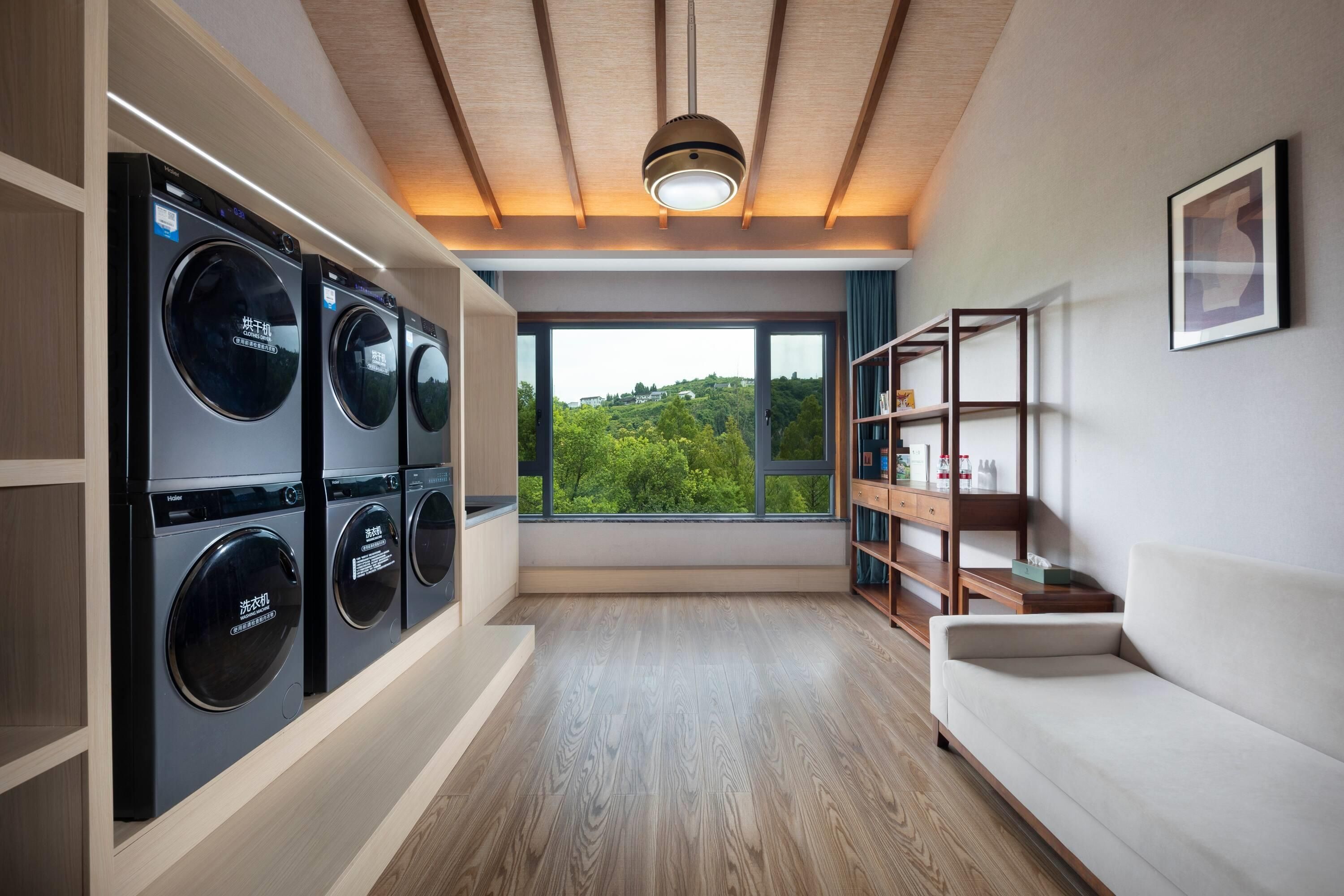 A laundry room with multiple washing machines, a wooden floor, a white sofa, and a large window with a view of greenery.